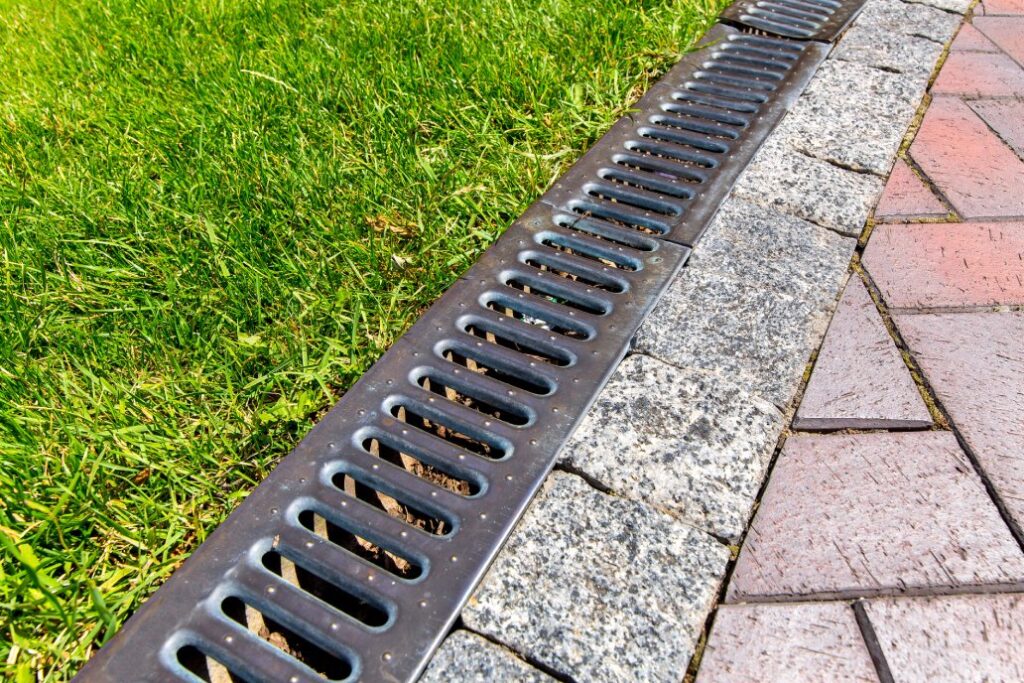 iron grate of a drainage system for storm water drainage from a pedestrian sidewalk near a green lawn - A Robinson Landscaping Services Drainage and Practical Solutions