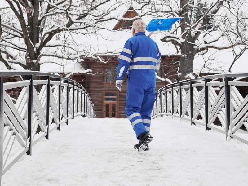 Elderly, handsome man with shovel on shoulder on winter bridge walking to wooden house. Concept of manual cleaning snow in winter after snow storm at yard.